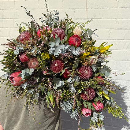 A bundle of Native Wildflowers featuring prominent pink and burgundy protea flowers, dusty pink protea blooms, green and silver eucalyptus foliage, and delicate filler stems, held by a person wearing a muted olive green shirt against a white painted brick wall.
