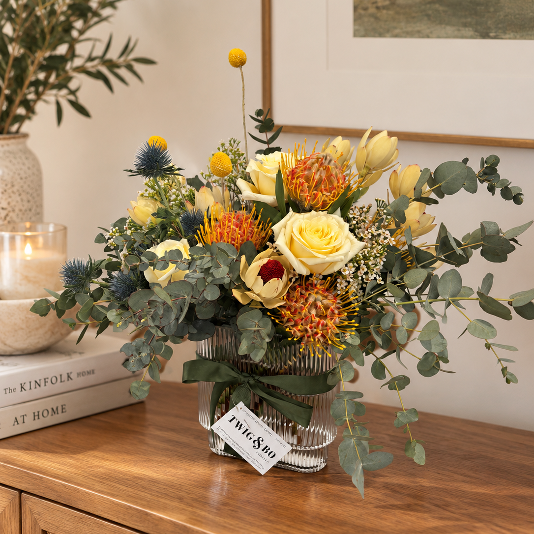 A floral arrangement in a ribbed glass vase on a wooden cabinet, featuring yellow roses, orange protea, blue thistles, and green eucalyptus foliages.